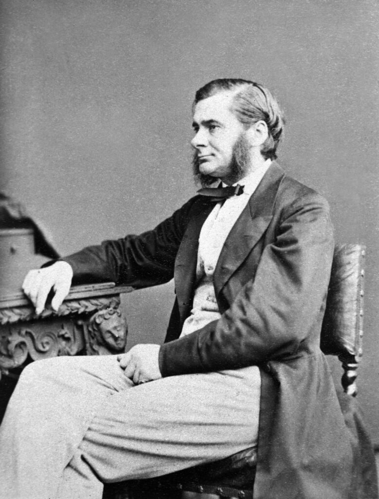Black and white photo of Thomas Henry Huxley, in Victorian times, sitting on a chair and wearing a jacket and bow tie. He has very impressive mutton chops.
