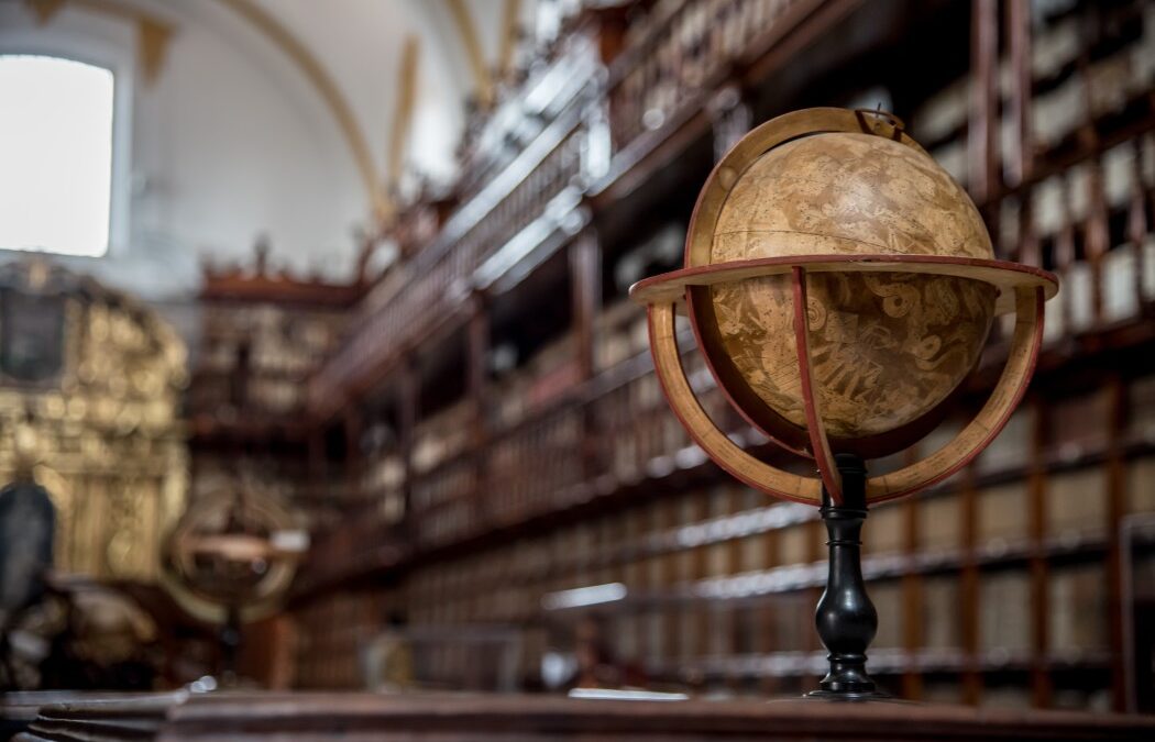 Featured image for Is Dark Academia Still Popular? to illustrate the dark academia aesthetic, it shows a picture of an antique library in the background with an old globe in focus on a table. The picture's vibe is dark and moody.