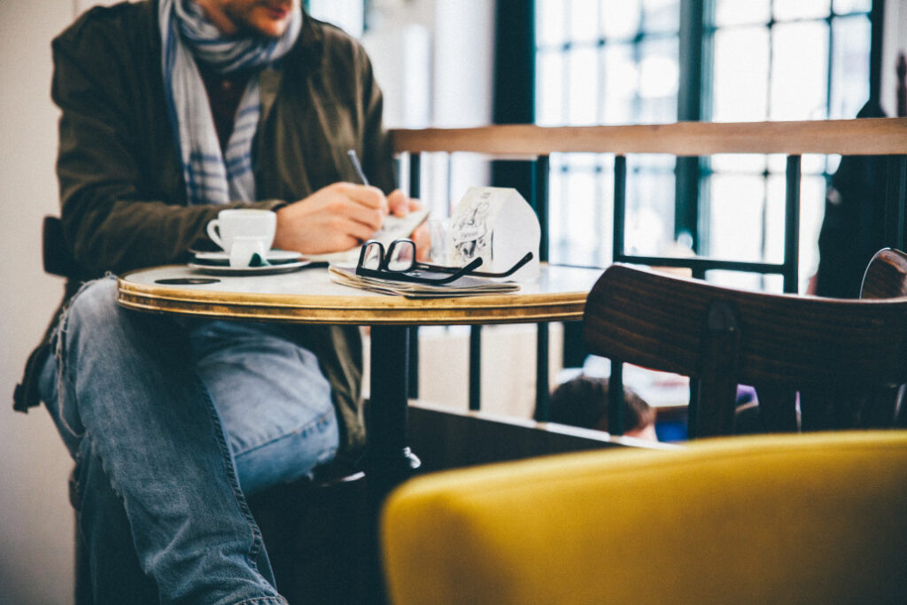 man writing in a notebook sitting at a cafe table- being a writer is one of the most common dark academia inspired jobs